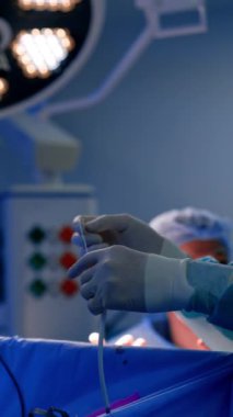 Lateks eldiven takan sıhhiyecilerin elleri operasyonda kullanılmak üzere cihazın kablolarını çözüyor. Arka planda ameliyat yapan maske ve şapkalı bir doktorun bulanık görüntüsü. Dikey video.