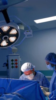 Beyaz erkek cerrah, kadın ameliyat hemşiresinin yardımıyla ameliyatta çalışıyor. Doktor, yuvarlak lambanın altındaki hastanın yanına oturur ve tavana asılır. Dikey video.