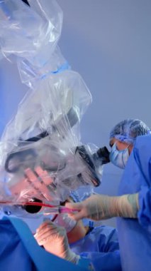 Kadın doktor mikroskoba bakar ve sıvıyı spreylemek için şırınga kullanır. Doktor ameliyatın yanında oturur. Nöroşirürji yapan doktorların görüş açısı düşük. Dikey video.