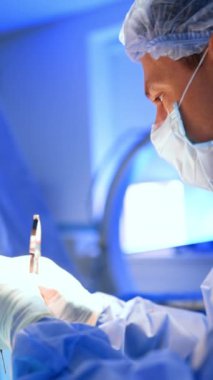 Metal forseps kullanan maskeli ve şapkalı bir erkek doktorun portresi. Yan görünüş yakın. Cerrah ameliyat ipliğini yana doğru uzatıyor. Dikey video.
