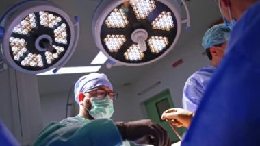 İki büyük yuvarlak lamba modern ameliyathanede faaliyet alanını aydınlatıyor. Bir grup doktor parlak ışık altında travma ameliyatı yapıyor..