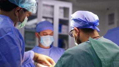 Operasyon masasını çevreleyen profesyonel tıbbi ekip. Sakallı cerrah iplik ve makas kullanır..