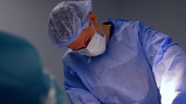 Maske, kep ve mavi bornoz giyen orta yaşlı beyaz erkek cerrah iş başında. Ameliyat sırasında bir doktorun portresi.