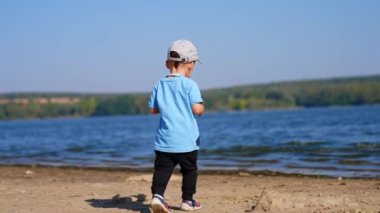Mavi tişörtlü küçük çocuk nehre doğru koşuyor. Küçük çocuk suya çakıl taşı atıyor. Bulanık arkaplan.