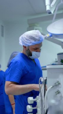 Mavi üniforma giyen beyaz sakallı doktor tıbbi ekipmanların başında duruyor. Gülümseyen kadın hemşire kameraya yaklaşıyor. Dikey video.