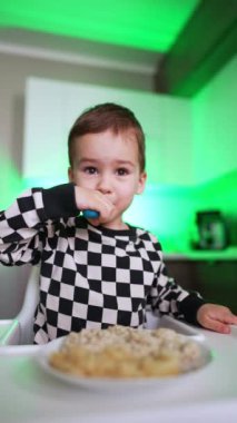 Kareli tişörtlü küçük pozitif çocuk yüksek sandalyede oturup yemek yiyor. Tatlı çocuk tabaktan yulaf lapası toplamak için çatal kullanıyor. Dikey video.