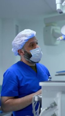 Maskeli, mavi üniformalı, sakallı, beyaz bir doktor ameliyat odasında elinde bir şişe solüsyonla dikiliyor. Sıhhiye operasyonel tiyatrodaki meslektaşlarıyla konuşuyor. Dikey video.