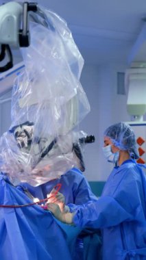 Sıhhiyeciler hastayı mikroskop altında inceliyor. Beyin cerrahı ve kadın doktorun ameliyatına yardım ediyor. Dikey video.