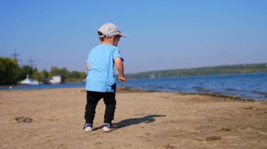 Bebek sahilde biraz kum topluyor. Çocuk nehre doğru yürür ve suya kum atar..