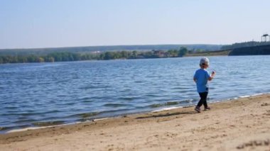 Küçük çocuk kumlu sahilde yürüyor. Küçük çocuk güneşli havalarda nehir kıyısında vakit geçiriyor..