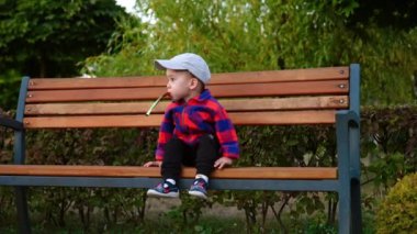 Şapkalı ve kareli tişörtlü güzel çocuk banktan iniyor. Bebek parktaki oyuncak kutusundan içiyor..