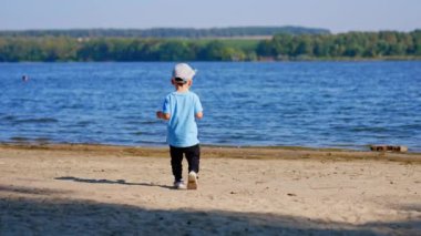 Nehre doğru giden küçük bir çocuğun arka görüntüsü. Mavi tişörtlü ve şapkalı çocuk suya kum atıyor..