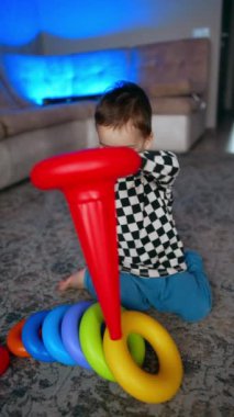 Genç erkek bebek yerde otururken renkli bir piramit tutuyor. Çocuk oyuncağı söker ve yeniden yapmaya başlar. Dikey video.