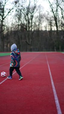 Genç erkek çocuk stadyumda topla oynuyor. Küçük bebek futbol oynamayı öğreniyor. Dikey video.