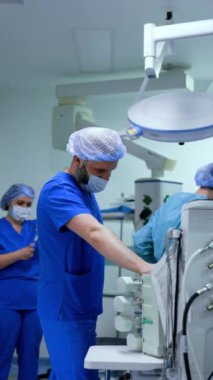 Mavi üniformalı sakallı doktor modern ameliyat odasındaki tıbbi ekipmanların yanında duruyor. Operasyonda çalışan bir anestezi uzmanı. Dikey video.