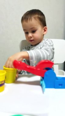 Masa başında oturan küçük beyaz bir çocuk plastik kalıplarla oynuyor. Mutlu çocuk evde kilden heykel yapıyor. Dikey video.