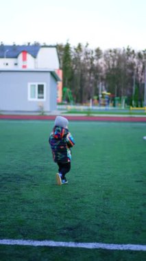 Sıcak giysili küçük bebek stadyuma koşuyor. Küçük çocuk topa tekme atarak futbol oynuyor. Dikey video.
