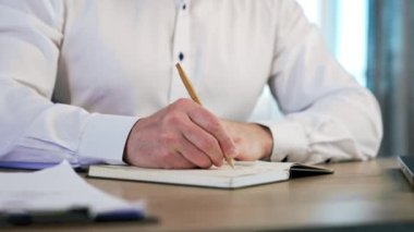 Beyaz gömlekli bir erkeğin elleri kâğıt deftere not alıyor. Bir tükenmez kalemle bir el yazısının görünümünü kapat.
