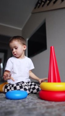 Tatlı bebek yerde oturup oyuncak piramidini parçalara ayırıyor. Sevimli çocuk oyun sayesinde şekilleri ve boyutları öğreniyor. Düşük açı. Dikey video.