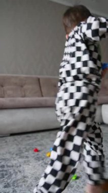 Evde damalı pijamalı neşeli bir çocuk oynuyor. Neşeli bebek oyuncakları toplayıp çekmeceye koyuyor. Dikey video.