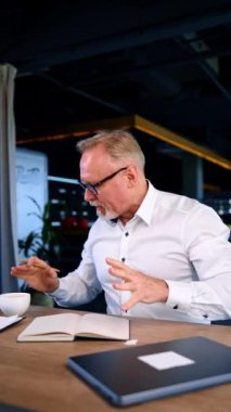 Ofisteki masasında oturan verimli çoklu görev sahibi iş adamı. Not alan, kahve içen ve enerjik hisseden bir adam. Dikey video.