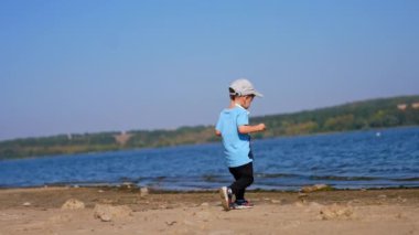 Mavi tişörtlü ve şapkalı bebek nehre doğru geliyor. Küçük çocuk taşları suya atıyor..