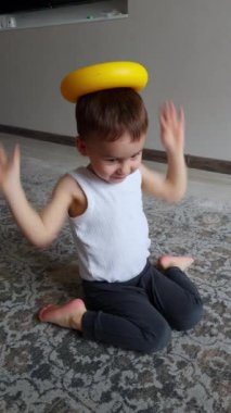 Evinde yerde oturan sevimli, beyaz bir bebek var. Çocuk kafasına oyuncak bir piramit yüzüğü takıyor ve düşürüyor, sonra da itiyor. Dikey video.