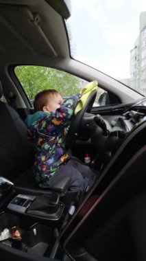 Babasının arabasının içini temizleyen kalın ceketli sevimli erkek bebek. Çocuk panellerin tozunu alırken sarı bezi kullanıyor. Dikey video.