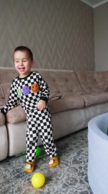 Evde damalı pijama giyen pozitif erkek bebek var. Neşeli çocuk odanın yanında topa vuruyor. Dikey video.