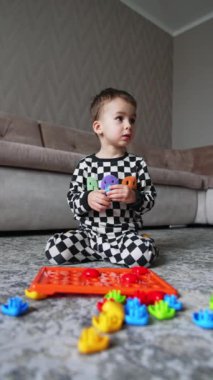 Kareli pijama giymiş beyaz bir çocuk yerde oturuyordu. Bebek bulmacayı çözer ve sevinçle sevinir. Düşük açı. Dikey video.