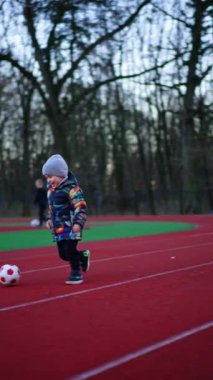 Mutlu gülümseyen çocuk stadyumun yanından geçen topu takip ediyor. Diğer büyük çocuklar arka planda bulanık olarak futbol oynuyorlar. Dikey video.