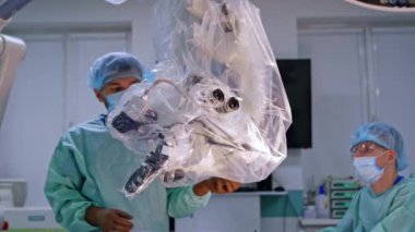 Erkek doktor mikroskobu ameliyathanede asılı tutuyor. Plastikle kaplı tıbbi ekipmanlar nöroşirürjde kullanılmak üzere hazırlanır..
