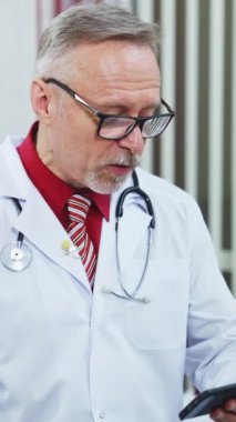 Kıdemli erkek doktor doktorun masasında oturuyor ve telefona bakıyor. Düşünceli deneyimli doktor video bağlantısı yoluyla hastaya danışır. Dikey video
