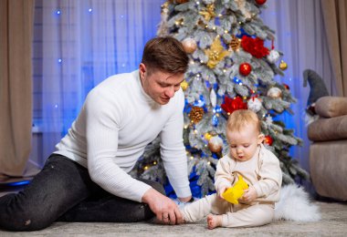 Baba ve bebek evdeki Noel ağacında birlikte oynuyorlar. Bir baba yerde oturuyor, bebeğiyle güzel dekore edilmiş bir Noel ağacının yanında.