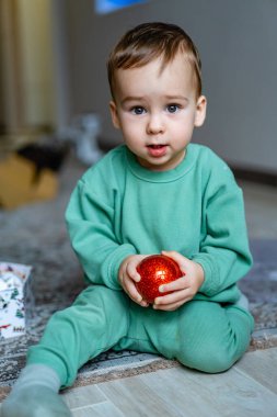 Küçük bir çocuk elinde kırmızı bir süs tutarak halının üzerinde oturuyor. Yeşil elbiseli küçük bir çocuk halıya oturmuş, parlak kırmızı bir süsü kavrayıp kameraya bakıyor..