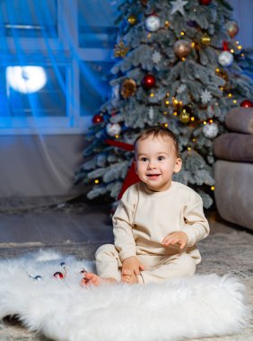 Bebek, kapalı bir Noel ağacı yakınındaki süslemelerle etkileşime giriyor. Küçük bir çocuk pofuduk bir paspasın üzerinde oturmuş, güzel dekore edilmiş bir Noel ağacının yanında süslemelerle oynuyor..