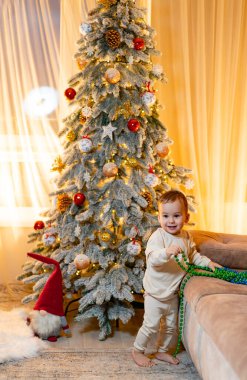 Mutlu bir bebek içeride neşeli bir Noel ağacının yanında oynar. Neşeli bir bebek süs ve ışıklarla süslenmiş bir Noel ağacının etrafında dolaşırken boncuk çeker..