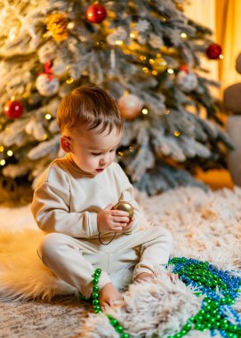 Bebek süslü bir Noel ağacının yanında süslemelerle oynuyor. Küçük bir çocuk yumuşak halıda oturur, güzel ışıklandırılmış bir Noel ağacının önündeki süsleri keşfeder.