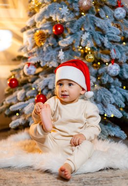 Noel ağacının yanında Noel Baba şapkası takan sevimli bir çocuk. Noel Baba şapkalı neşeli bir çocuk güzel dekore edilmiş bir Noel ağacının önünde pofuduk bir halıya oturur.