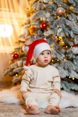 Evdeki süslü Noel ağacının yanında neşeli şapkalı şirin çocuk. Küçük bir çocuk yumuşak bir halıya oturur, Noel Baba şapkası takar, yakınlardaki güzel dekore edilmiş Noel ağacına merakla bakar..