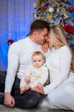 Evde Noel ağacı süsleyerek bebekle aile kutlaması. Neşeli bir aile anı, güzel dekore edilmiş bir Noel ağacının yanında bebeğiyle ebeveynleri ağırlıyor.