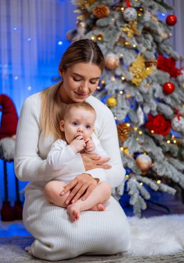 Anne ve bebek Noel 'i süslü bir Noel ağacında kutluyor. Bir anne bebeğini nazikçe güzel süslenmiş bir Noel ağacının yanında, şenlik ortamında tutar.