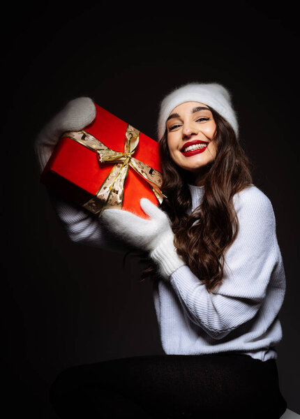 Smiling woman in winter attire holding a festive gift box at night. A joyful woman in cozy winter clothing happily presents a beautifully wrapped gift
