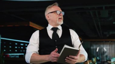 Kendine güvenen yetişkin iş adamı beyaz gömlek ve ofiste kumlu siyah yelek giyiyor. Elinde not defteri olan adam düşük açı görünümlü..