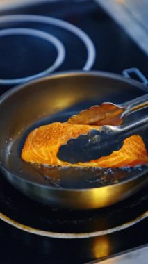 Metal maşalar somon parçasını kızgın yağlı tavaya koyar. Fırında balık pişiriyorum. Kapatın. Dikey video.