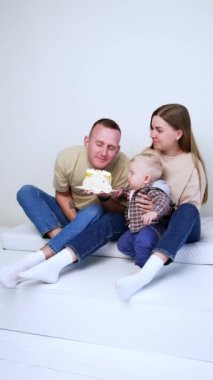 Duvarda yerde oturan üç kişilik genç bir aile. Küçük çocuk babasının elinde tuttuğu ve kokladığı pastaya bakıyor. Dikey video.