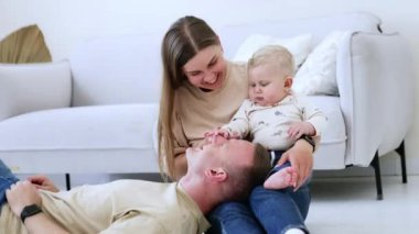 Adam karısının dizlerinin üstüne çöktü. Annenin elinde oturan sarışın çocuk babasının burnuna dokunuyor. Oda arkaplanı.