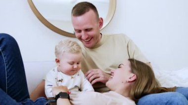 Üç kişilik sevimli genç bir aile evdeki kanepede yatıyor. Kadın erkeğin kucağına yatar ve erkek bebek annesinin saçına dokunur. Düşük açı görünümü.