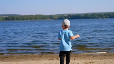 Sahilde denize doğru koşan bir çocuğun arka görüntüsü. Yürüyen çocuk durur ve taşı nehre atar..