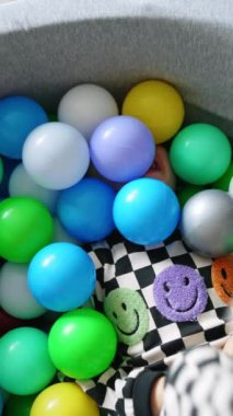 Küçük bebek plastik toplarla kaplı kuru bir havuzda yatıyor. Mutlu çocuk mutlu bir şekilde oturur. Üst Manzara. Dikey video.